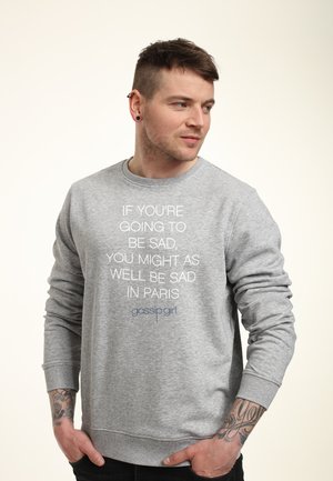 Mand med tatoveringer iført grå sweatshirt med teksten "Hvis du skal være trist, kan du lige så godt være trist i Paris" står med hænderne i lommerne.