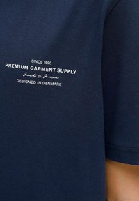 Námořnická modrá tričko s bílým textem: "Od roku 1990 Prémiová oděvní zásoba Jack & Jones Navrženo v Dánsku" na přední straně.