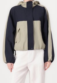 Färgsorterad vindjacka i marinblått och ljus olivgrönt. Har en huva, elastiska ärmslut och logotypdetalj på bröstet. Lättviktsmaterial.