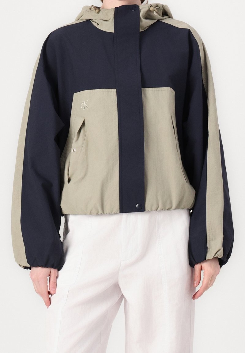 Färgsorterad vindjacka i marinblått och ljus olivgrönt. Har en huva, elastiska ärmslut och logotypdetalj på bröstet. Lättviktsmaterial.