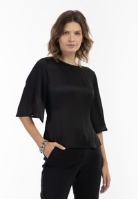 Bluză neagră, semi-transparentă, cu guler rotund, mâneci evazate până la trei sferturi și textură netedă, purtată cu pantaloni negri. Brățară argintie pe încheietura mâinii.
