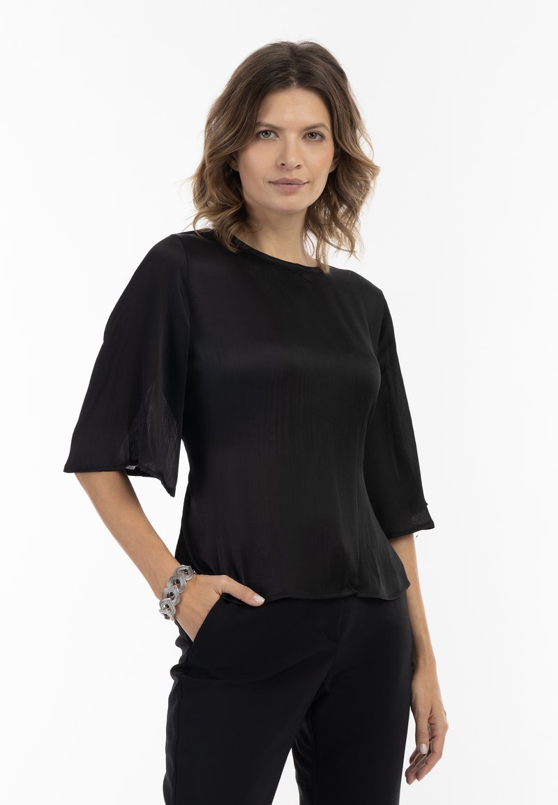 Bluză neagră, semi-transparentă, cu guler rotund, mâneci evazate până la trei sferturi și textură netedă, purtată cu pantaloni negri. Brățară argintie pe încheietura mâinii.