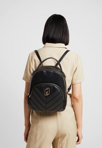 Mochila de cuero negro con un diseño en chevron y pequeños remaches. Presenta un logo dorado en la parte frontal y correas dobles para los hombros.