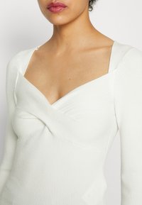 Haut blanc à manches longues côtelées avec un col en V profond et un détail torsad é à l'avant. Présente une coupe ajustée et une texture lisse.