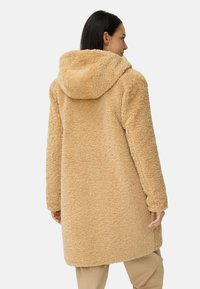 Beige, fluffy jas met een capuchon, met een textuur van een zachte buitenkant en een relaxte pasvorm. Het ontwerp is knielang met lange mouwen.