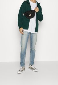 Green zip-up hoodie over a white t-shirt, paired with light blue jeans and black-and-white zebra print sneakers. A black fanny pack is worn.