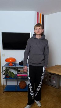 Νεαρός άνδρας με γκρι φούτερ Timberland και μαύρο παντελόνι Adidas στέκεται στο σαλόνι με πορτοκαλί λάμπα, μπάλα του μπάσκετ και τηλεόραση στον τοίχο.