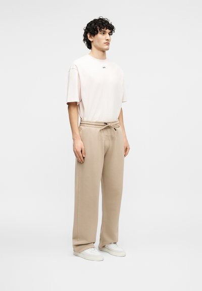 Νέος άνδρας στέκεται φορώντας ένα ανοιχτό ροζ φαρδύ T-shirt, μπεζ άνετη φόρμα με κορδόνι και λευκά αθλητικά παπούτσια σε απλό φόντο.