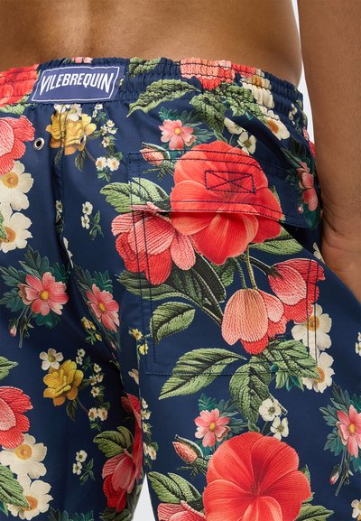 Gros plan sur un short de bain bleu marine avec un imprimé floral multicolore, une poche arrière et une étiquette de marque Vilebrequin sur la ceinture.