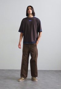 Loose-fit dark t-shirt with a round neck and blue accents, paired with brown wide-leg pants and beige sneakers on a gray surface.