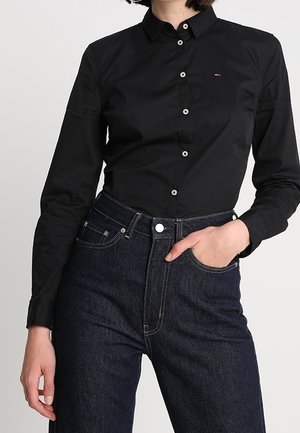 Person iført en tætsiddende sort skjorte med knapper og mørkeblå jeans med høj talje, med den ene hånd i en forlom.
