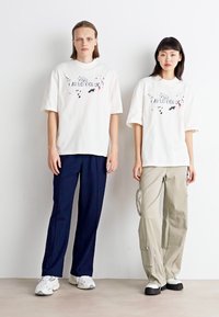 Hvide grafiske t-shirts med farverige designs, parret med navy og beige løstsiddende bukser; teksturerede sneakers fuldender outfittet.