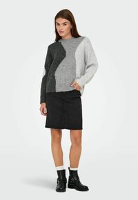 Femme portant un pull gris à blocs de couleur, une jupe en denim noir, des chaussettes blanches et des bottines noires, se tenant avec une main dans la poche.