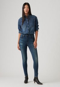 Žena stojaca oblečená v modrej denimskej košeli zastrčenej do nohavíc s vysokým pásom a čiernych členkových čižmách, na jednofarebnom pozadí.