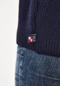 Marineblauer Strickpullover mit geripptem Muster, versehen mit einem kleinen Logoetikett in Rot und Weiß, kombiniert mit verwaschenen blauen Jeans.