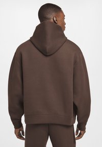 Brun hættejakke med en stor, rund snorhætte, løs pasform og ribbede manchetter. Glat tekstur og enkel design uden mønstre.