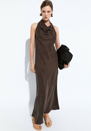 Donna che indossa un abito lungo senza maniche marrone scuro, tiene in mano una pochette nera intrecciata e indossa sandali beige piatti su sfondo bianco.