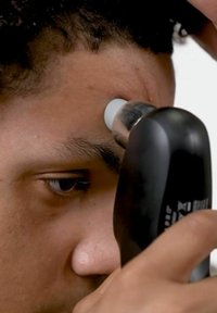 Tondeuse à cheveux noire avec une pointe blanche tenue près de la peau, se concentrant sur la zone du front. Vue en gros plan de l'œil d'une personne et de la texture de sa peau.