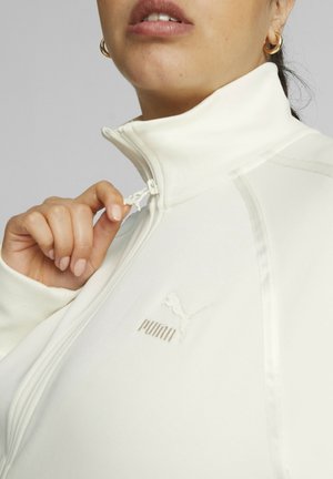 Veste de sport blanche à col haut, fermeture éclair, et logo Puma discret. Le tissu présente une texture lisse et un design minimaliste.