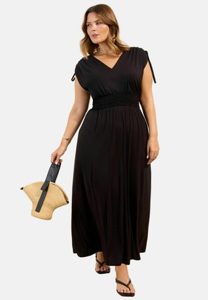 Frau trägt ein schwarzes ärmelloses Maxikleid mit geraffter Taille, hält eine gewebte Handtasche mit schwarzen Trägern und trägt schwarze Sandalen.