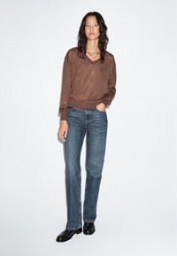 Kvinna som står mot en vit vägg iförd en brun V-ringad tröja, blå raka jeans, svarta skor och ett silverkedja halsband med handen i fickan.