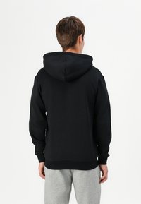Sudadera negra con una gran capucha, mangas largas y puños y dobladillo de canalé. Combinada con pantalones de chándal grises, mostrando un look casual.