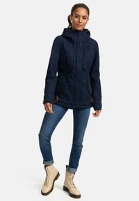 Navy hooded jacket with a textured design, adjustable drawstrings, two front pockets, and buttons. Paired with blue jeans and beige lace-up boots.