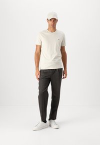Tricou din bumbac bej cu un logo mic, pantaloni negri închis și adidași albi. O șapcă de culoare deschisă completează ținuta. Design simplu și casual.