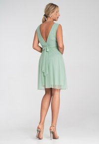 Mintgrünes Kleid mit V-Ausschnitt, tailliert mit einer Schleife gebunden und knielangem, durchsichtigem Rock. Model trägt klare Absätze.
