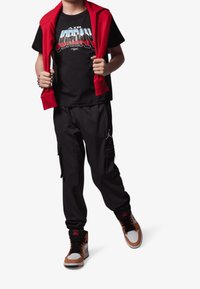 Czarne spodnie cargo, czarna koszulka z grafiką 'AIR JORDAN', czerwona bluza zarzucona na ramionach i brązowe/białe sportowe buty.