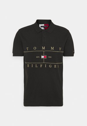 Polo noir à manches courtes avec col, texte jaune "Tommy Hilfiger", logo central et "EST. 1985" devant.