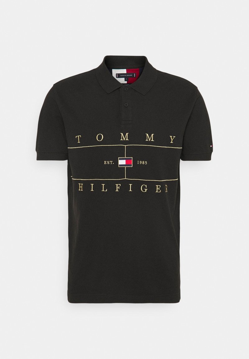 Polo noir à manches courtes avec col, texte jaune "Tommy Hilfiger", logo central et "EST. 1985" devant.