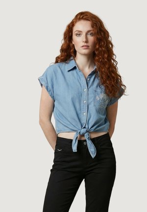 Femme aux longs cheveux roux bouclés, portant une chemise en denim bleu clair à manches courtes nouée et un pantalon noir, debout devant un fond uni.