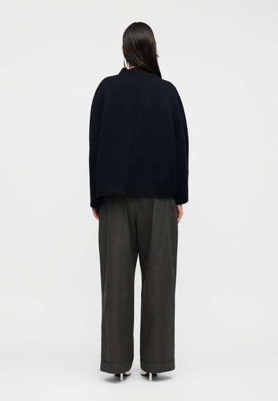 Ανοιχτή μπλούζα σε σκούρο ναυτικό μπλε με στρογγυλή λαιμόκοψη, άνετη εφαρμογή και χοντρή υφή, συνδυασμένη με φαρδιά γκρι παντελόνια.