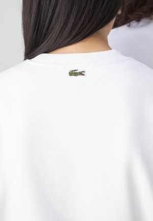 Personne aux longs cheveux foncés portant une chemise blanche avec un petit logo crocodile vert dans le haut du dos près de l'encolure.