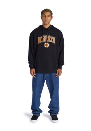 Sudadera negra con el logo "DC Shoe Co" en naranja y blanco. Jeans azules holgados y zapatillas negras. Cuenta con un bolsillo frontal y una capucha ajustable.