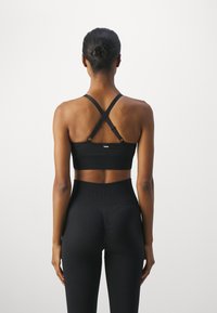 Sutiã desportivo preto com textura canelada e alças cruzadas nas costas. Usado com leggings pretas de cintura alta, apresentando um logótipo da marca na cintura.