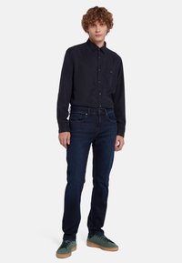 Camicia bottone di un blu scuro con una tasca sul petto, abbinata a jeans blu scuro aderenti e sneakers verdi con suola marrone chiaro.