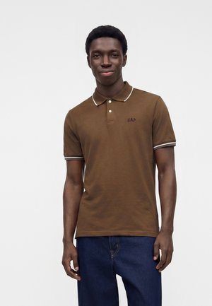 Mand står mod en ensfarvet baggrund iført en brun GAP-polo med hvid kant og blå jeans, kigger mod kameraet.