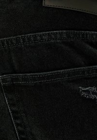 Černá džínová tkanina s viditelným šitím, obsahující malou opotřebovanou oblast a kapsu. Textura působí pevně a mírně vybledle.