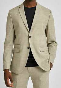 Ljusbeige blazer med två framfickor och en enkel knappstängning, tillsammans med en svart skjorta. Slätt, texturerat tyg.