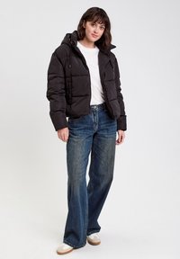 Doudoune noire avec capuche, dotée d'une fermeture éclair et d'un design matelassé. Portée avec un jean large bleu clair et des chaussures décontractées beiges.