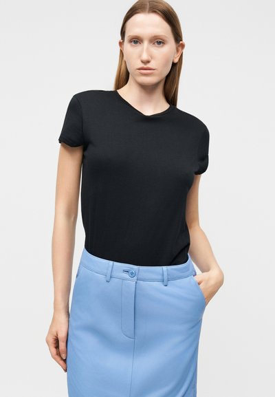 Γυναίκα με ίσια ανοιχτό καφέ μαλλιά, που φοράει μαύρο μπλουζάκι με κοντά μανίκια, το οποίο είναι μπαλωμένο μέσα σε μια ψηλόμεση ανοιχτό μπλε φούστα με τσέπες.