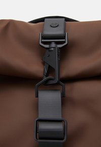 Hnedá vodotesná taška s čiernym nastaviteľným popruhom a detailom uzáveru z čierneho kovu, s textúrovaným povrchom pre zvýšenú odolnosť.