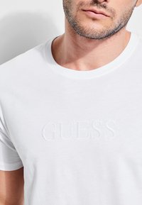Valge puuvillane T-särk, millel on ümar kaelus ja reliefne "GUESS" logo esiküljel, esitleb sujuvat tekstuuri ja juhuslikku disaini.