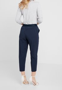 Pantalon bleu marine à coupe fuselée, taille élastique et deux poches arrière. Associé à une chemise à manches longues gris clair et des escarpins blancs.