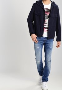 Marinblå lättviktsjacka med huva, två framfickor, bärs över en vit t-shirt med tryck, tillsammans med slitna blå jeans och vita sneakers.