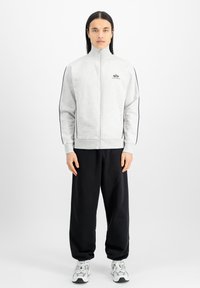 Hellegrauer Zip-Hoodie mit hohem Kragen, schwarzen Seitenstreifen und Logodetail auf der Brust, kombiniert mit schwarzen lässigen Hosen und Sneakern.