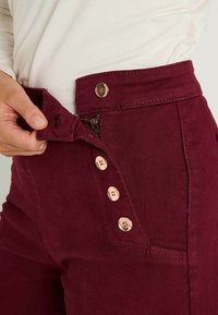 Pantalones de tiro alto burdeos con cremallera y cierre de cinco botones. Presenta botones de tono cobre y una tela de algodón texturizada.