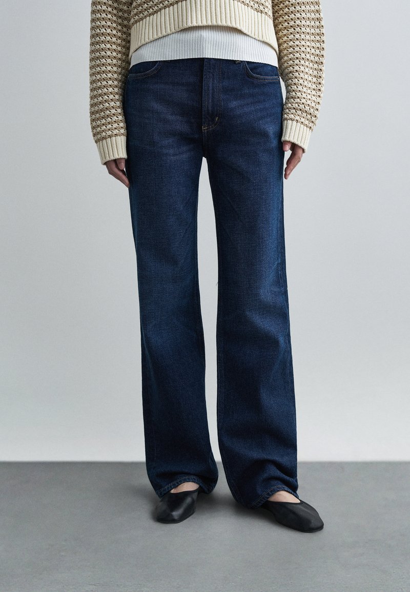 Džínsy tmavomodrej farby s širokými nohavicami, hladkou textúrou a klasickým dizajnom s piatimi vreckami, skombinované s čiernymi plochými topánkami a pleteným svetrom.